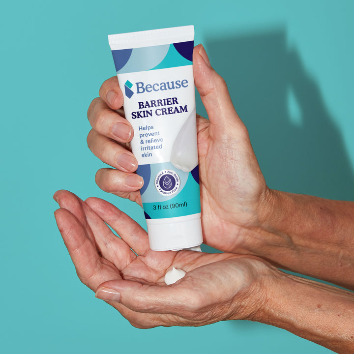Hand holding a tube of 'Because' Barrier Skin Cream against a teal background