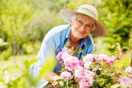 Senior adult woman wearing a brown sun hat and light blue shirt kneeling beside a flower bed in the garden