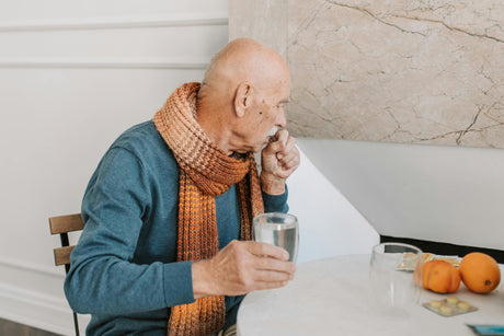 Older adult male wearing a long sleeved blue shirt and orange scarf, sitting at a small round table with oranges and medication on the surface, holding a glass of water