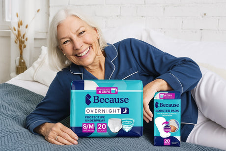 Older adult woman with white, chin-length hair wearing blue shirt and white pants lying on her side on a bed with Because Overnight Underwear product package and Because Booster Pad package showcased.