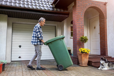 Older man rolling trash can in front of house with husky lying near the front porch