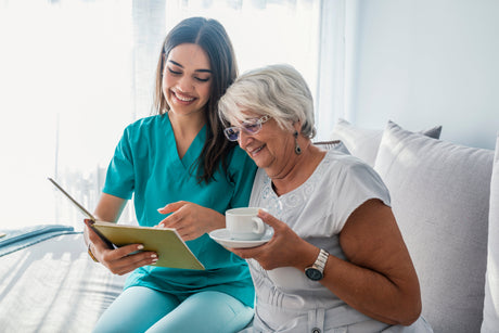 An older woman holding a cup of tea and her caregiver smiling as they review a notebook while sitting on a bed in a brightly lit bedroom 