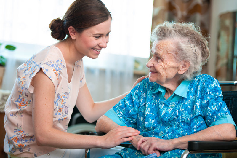 A young woman and older adult woman sitting beside each other smiling. The young woman has her hands resting on the arm and shoulder of the older woman. Room blurred in background.