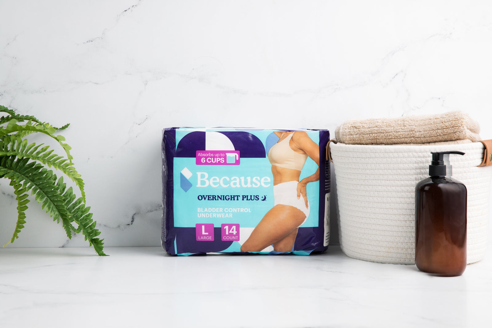 Because Overnight Plus Underwear product package set in center of marble counter beside a fern to the left and a small hamper filled with beige towels and a brown bottle of soap to the right