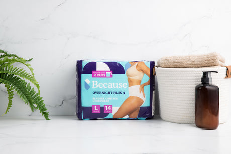 Because Overnight Plus Underwear product package set in center of marble counter beside a fern to the left and a small hamper filled with beige towels and a brown bottle of soap to the right