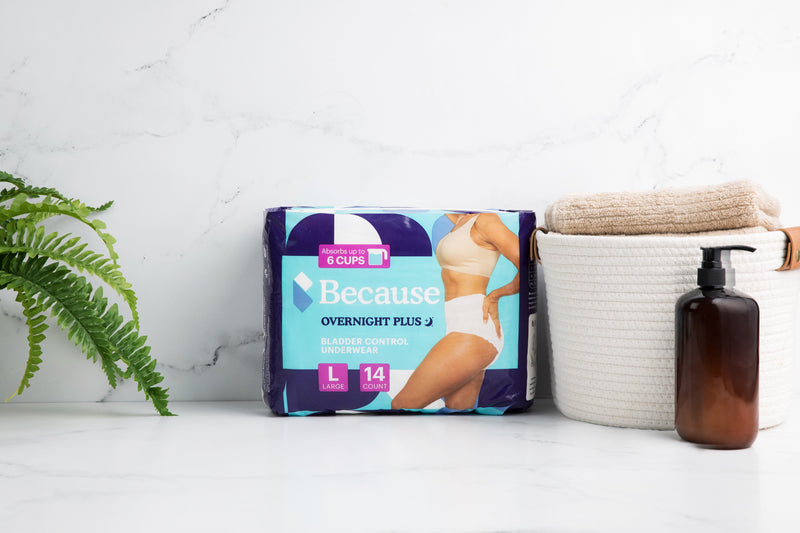 Because Overnight Plus Underwear product package set in center of marble counter beside a fern to the left and a small hamper filled with beige towels and a brown bottle of soap to the right