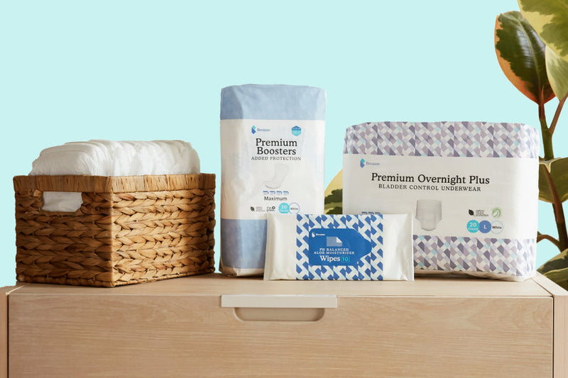 Packages of Because Overnight Underwear, Because Cleansing Wipes, and Because Booster pads sitting on top of a dresser with a basket filled with Because Overnight Underwear. Plant in background against blue wall 