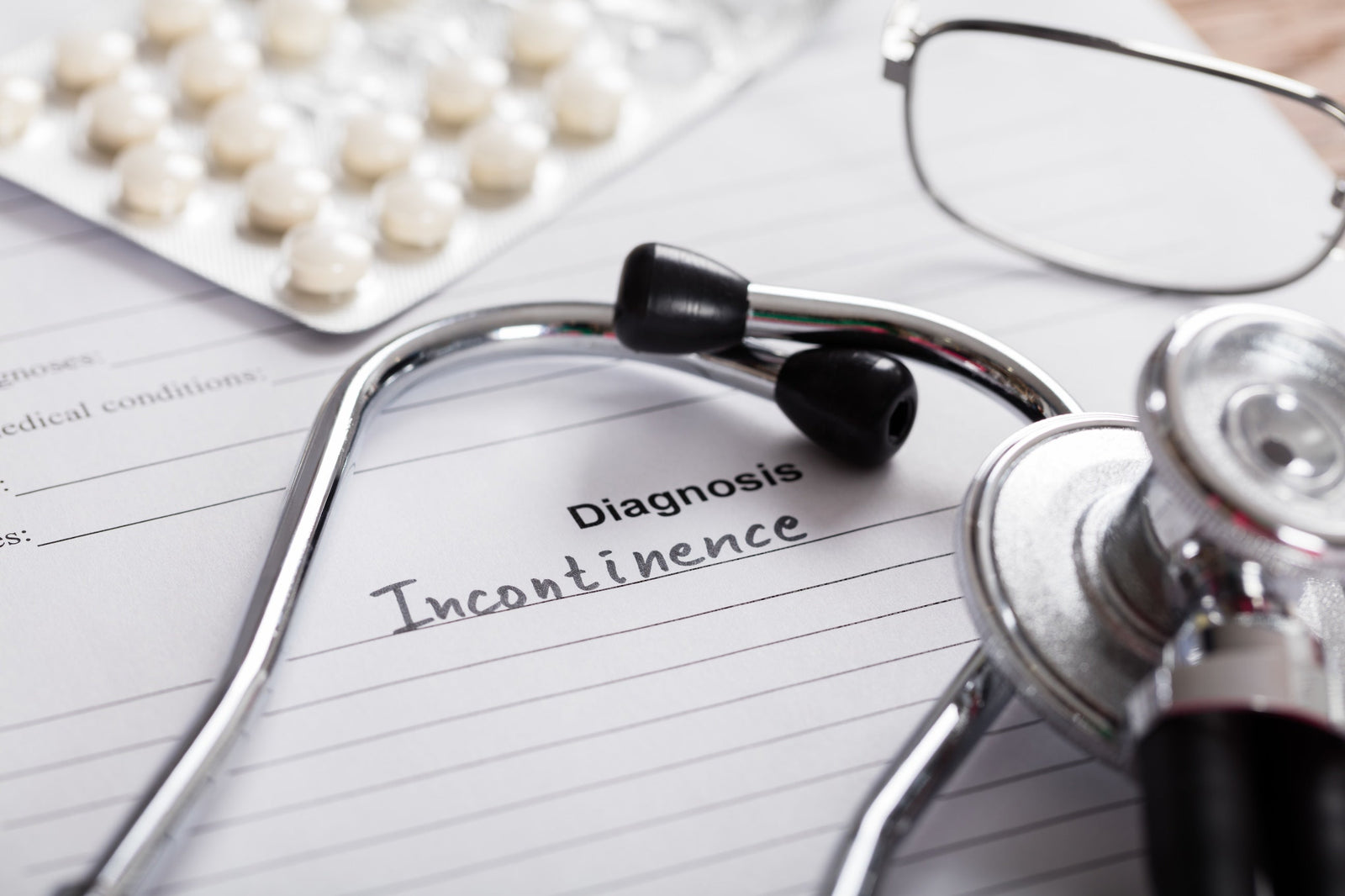 Diagnosis paper with "incontinence" written on a line, with medication packet, stethoscope, and glasses arranged on top.