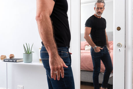 Man with glasses wearing dark jeans and black t-shirt looking in the mirror in a bedroom.