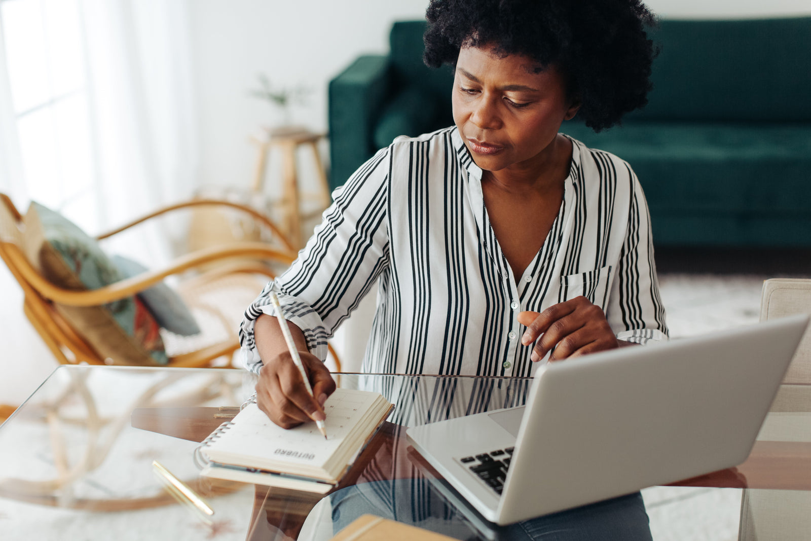 African American woman sitting at glass table with open laptop, writing in a journal with arm chair and sofa blurred in background.
