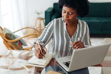 African American woman sitting at glass table with open laptop, writing in a journal with arm chair and sofa blurred in background.
