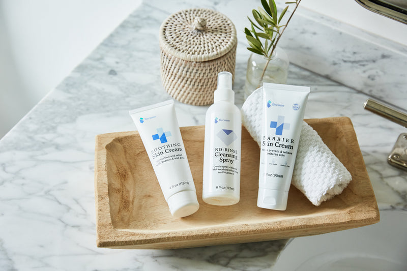 Because skin cream, no-rise cleansing pray, and barrier cream products on wooden tray resting on marble bathroom counter.