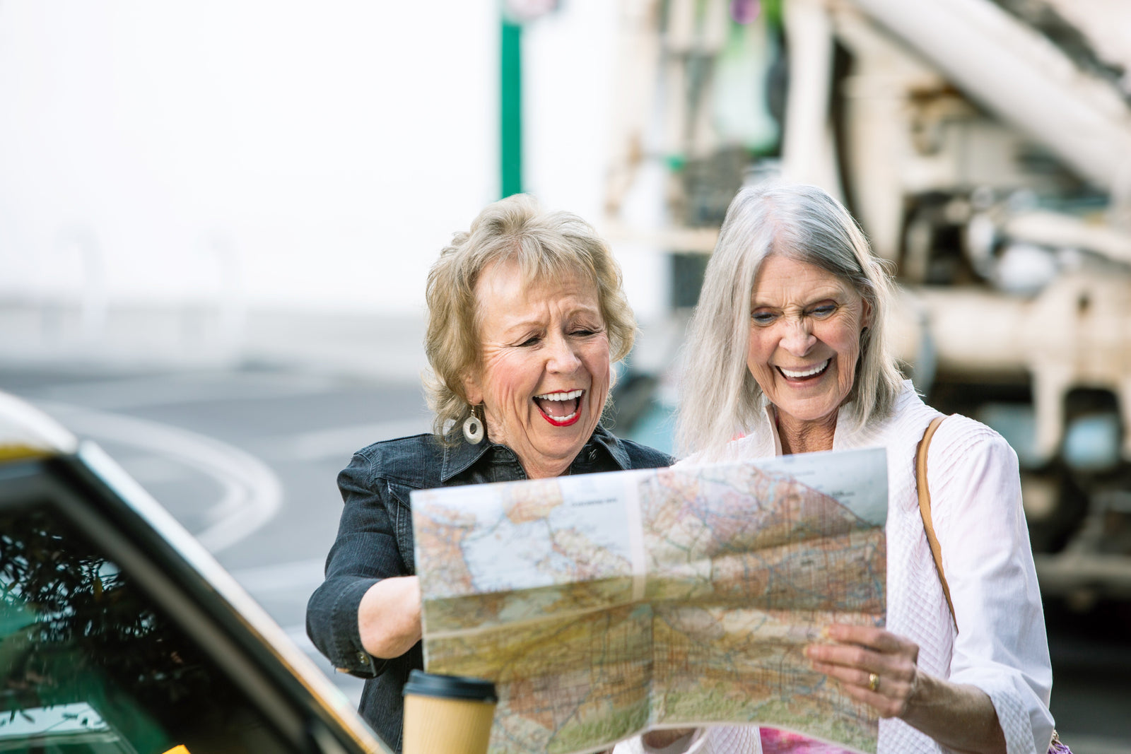Two senior adult women standing outside of a car laughing together as they looks at a map