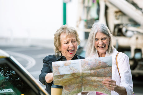 Two senior adult women standing outside of a car laughing together as they looks at a map