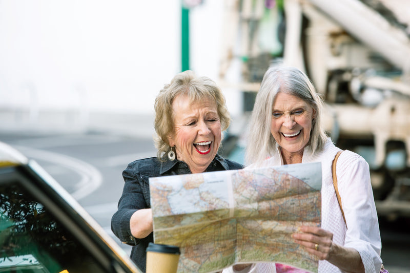 Two senior adult women standing outside of a car laughing together as they looks at a map