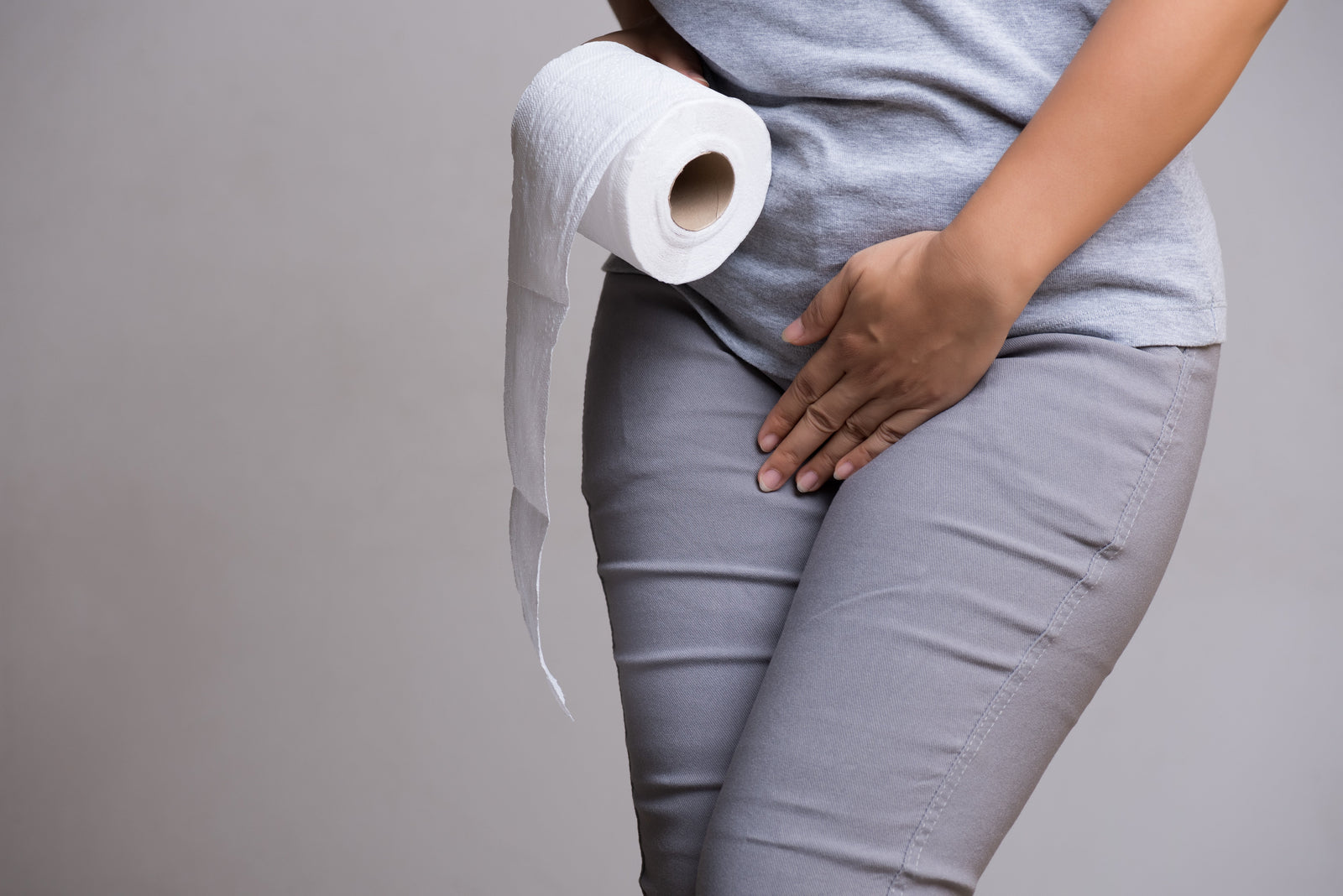 Woman in grey jeans and light grey t-shirt pressing a hand over stomach and the other hand holding a roll of toilet paper on a dark grey background