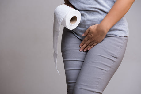 Woman in grey jeans and light grey t-shirt pressing a hand over stomach and the other hand holding a roll of toilet paper on a dark grey background