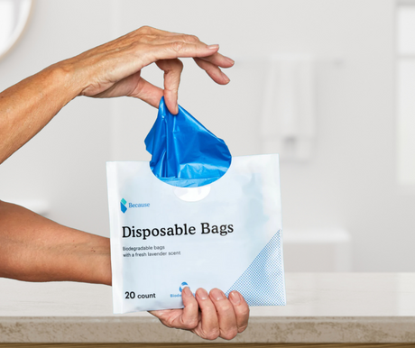 A pair of hands holding a package of Because Disposable Bags and pulling product from the package with blurred bathroom in the background