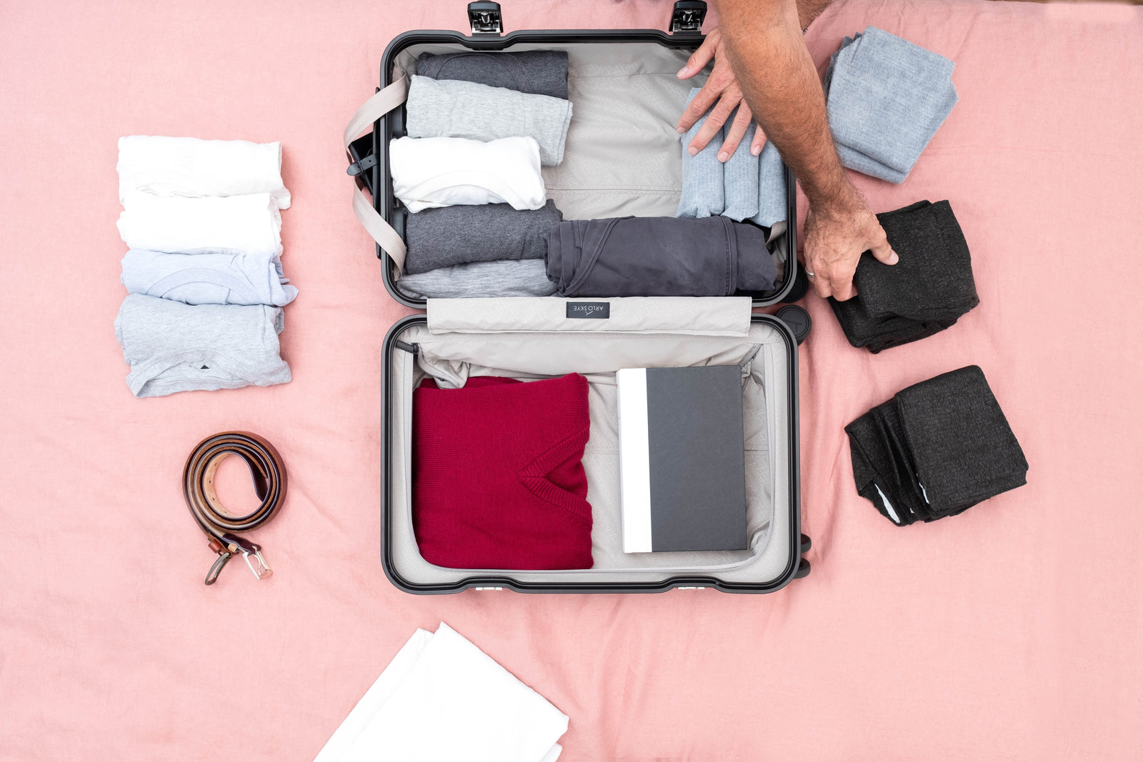 An opened suitcase on a salmon-colored bedspread with folded clothes beside and inside suitcase with hands packing folded adult underwear in