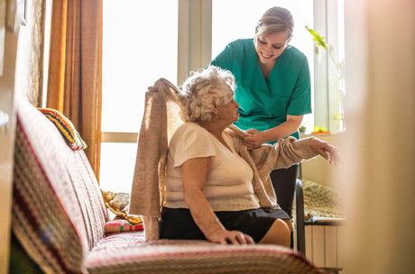 A caregiver in a green shirt helping an older adult get from her wheelchair onto her bed