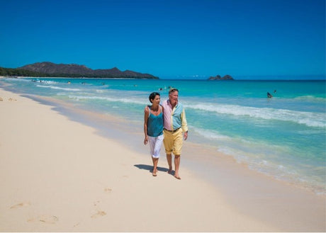 An older couple walking down a sandy beach on a bright blue sky day