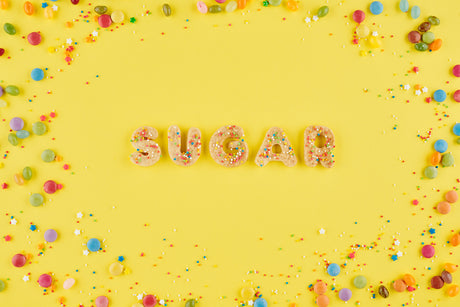 Lettered cookies spelling out the word 'sugar' sit on a purple background surrounded by sprinkles.