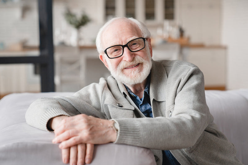 Smiling happy healthy caucasian senior old elderly man grandfather relaxing resting on the sofa couch looking at camera with toothy smile at home.