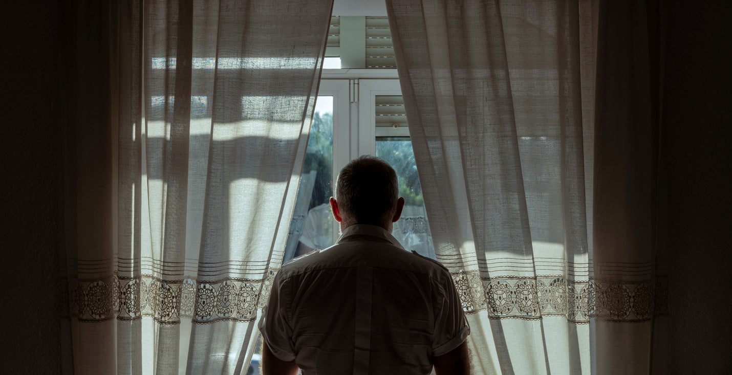 A man looks out the window from a dark room.