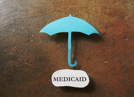 A paper umbrella over a cut out of the word "medicaid".