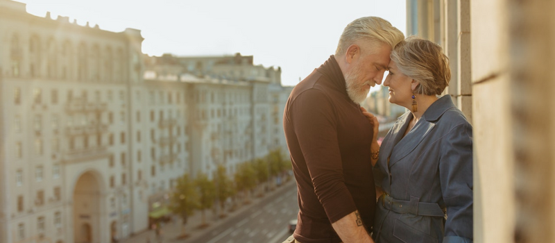 A man and a woman stand on a balcony in an old city with their heads pressed together lovingly.