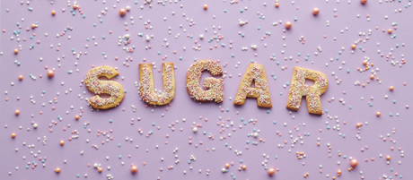 Lettered cookies spelling out the word 'sugar' sit on a purple background surrounded by sprinkles.