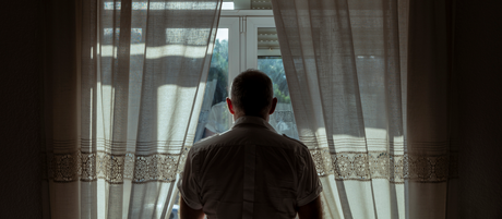 A man looks out the window from a dark room.