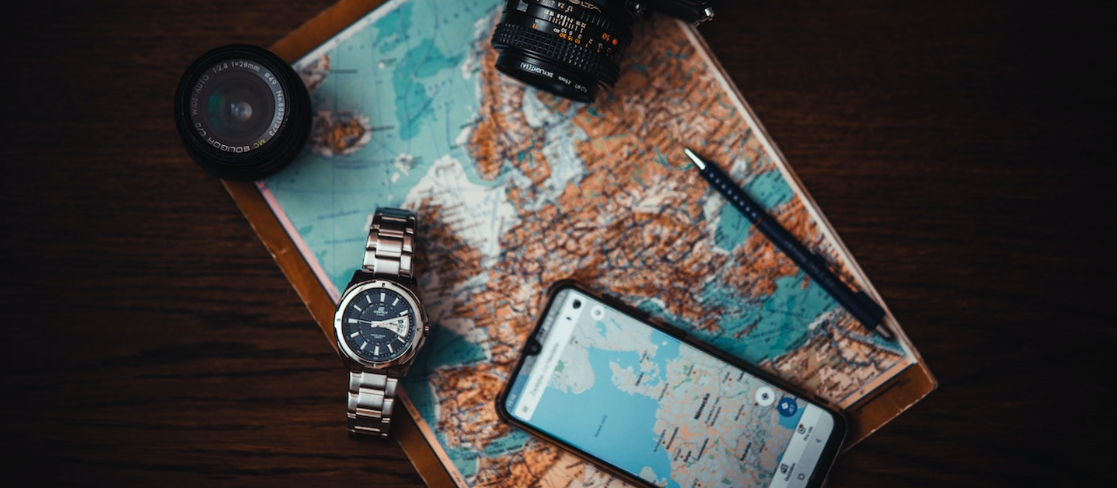 A printed map displaying Europe with a smartphone, pen, watch, DSLR camera and lens, and a pen laid on it.