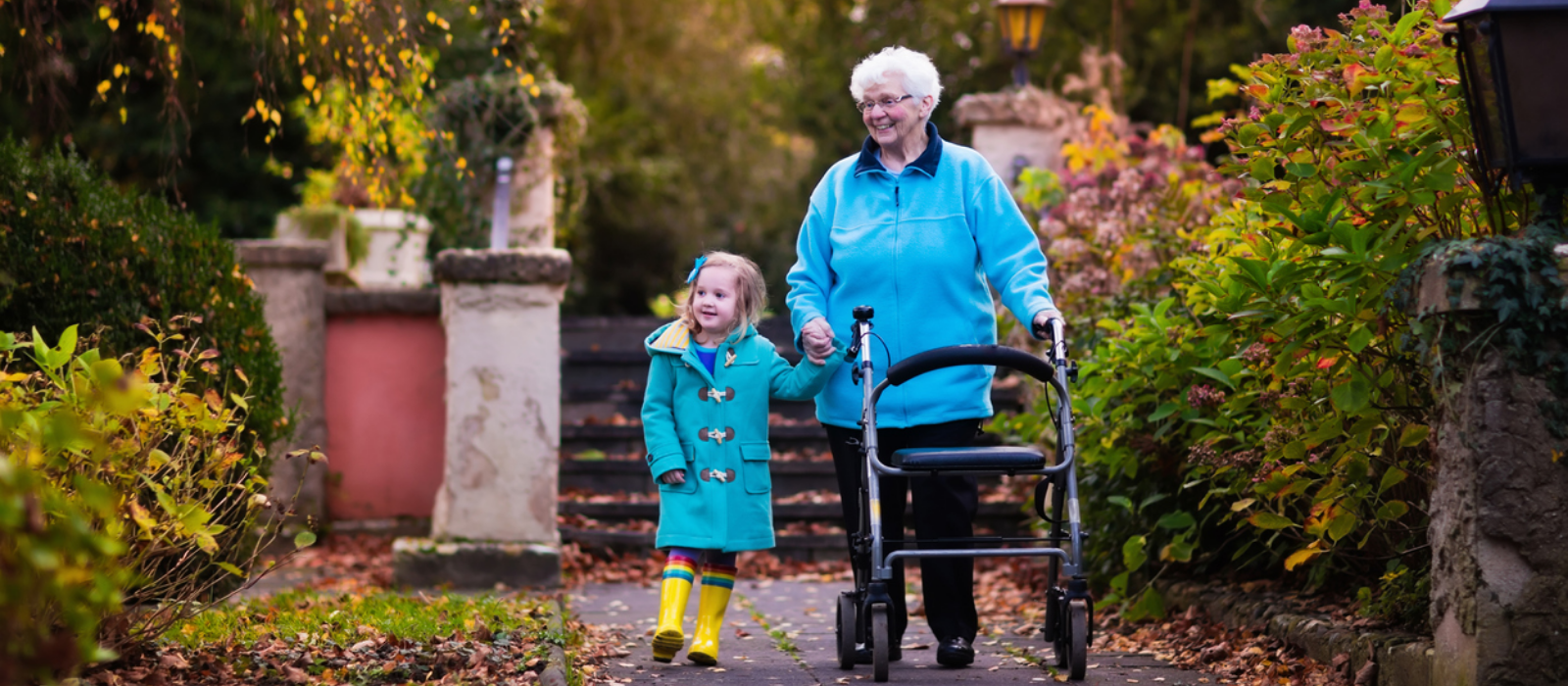 A grandmother with a rollator and her granddaughter walk down a park path smiling.