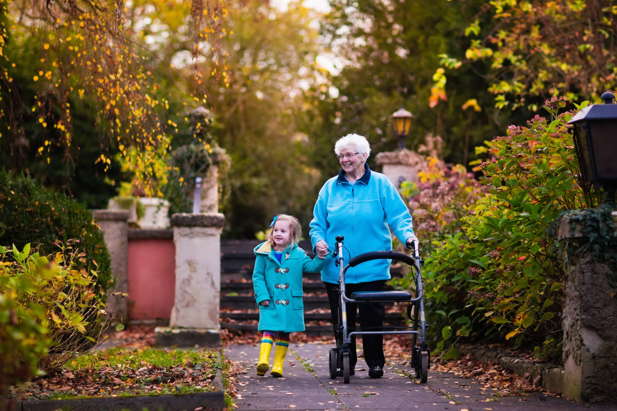 A grandmother with a rollator and her granddaughter walk down a park path smiling.