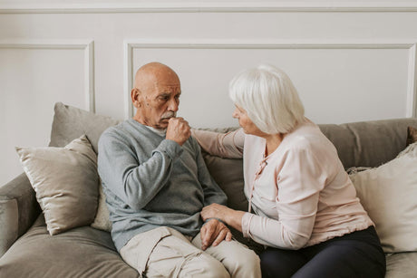 A man sitting on a couch coughs while a woman comforts him.