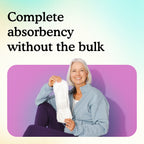 A smiling older woman with white hair sits against a purple wall holding up a slim Maximum pad, showing full absorbency without bulk.