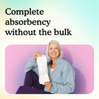 A smiling older woman with white hair sits against a pink wall holding up a slim Moderate pad, showing full absorbency without bulk.