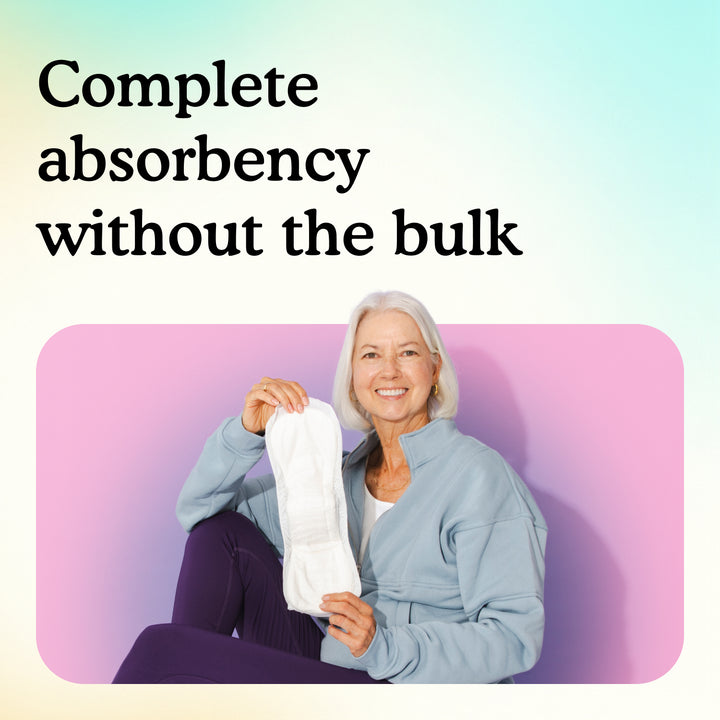 A smiling older woman with white hair sits against a pink wall holding up a slim Moderate pad, showing full absorbency without bulk.
