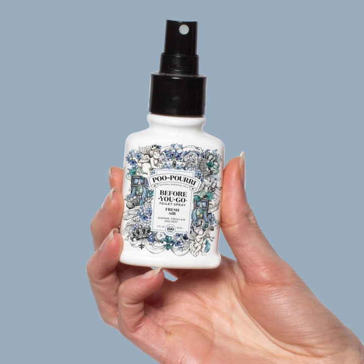 Hand holding a Poo-Pourri bottle with decorative label on a light blue background