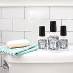 Three Poo-Pourri bottles sitting on a toilet tank lid beside a washcloth and spray bottle with a tile background