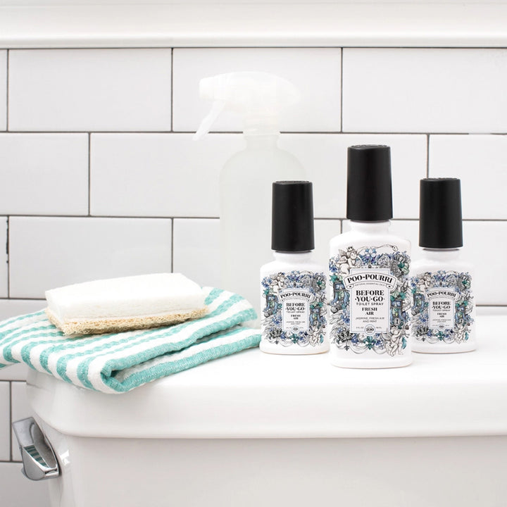 Three Poo-Pourri bottles sitting on a toilet tank lid beside a washcloth and spray bottle with a tile background