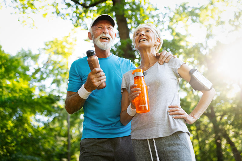 A man and woman stand in the woods after working out smiling with water bottles.